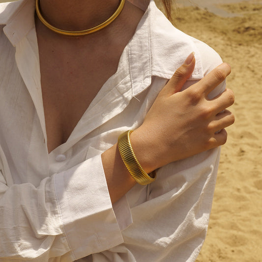 Aurora Luxe armband in gouden kleur, gecombineerd met een verfijnde uitstraling, gedragen met een witte blouse. Het tijdloze ontwerp van de armband, met een elegante structuur, straalt luxe en stijl uit, en is perfect voor elke gelegenheid.